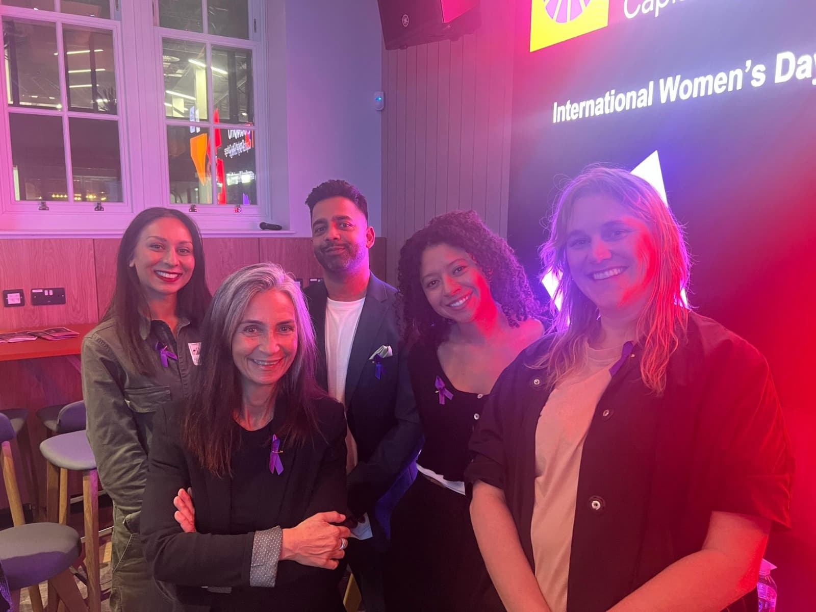 Five panellists at an International Women's Day event, smiling under stage lights.