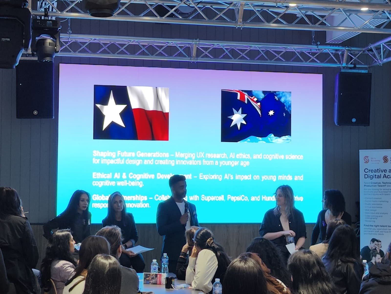A speaker presenting to a seated audience, with Texas and Australian flags on the screen behind.