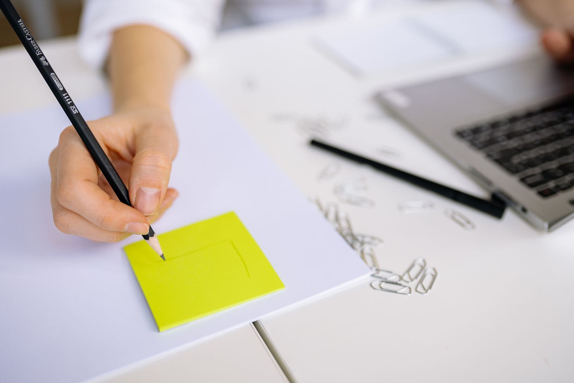 A hand sketching on a sticky note beside a laptop.