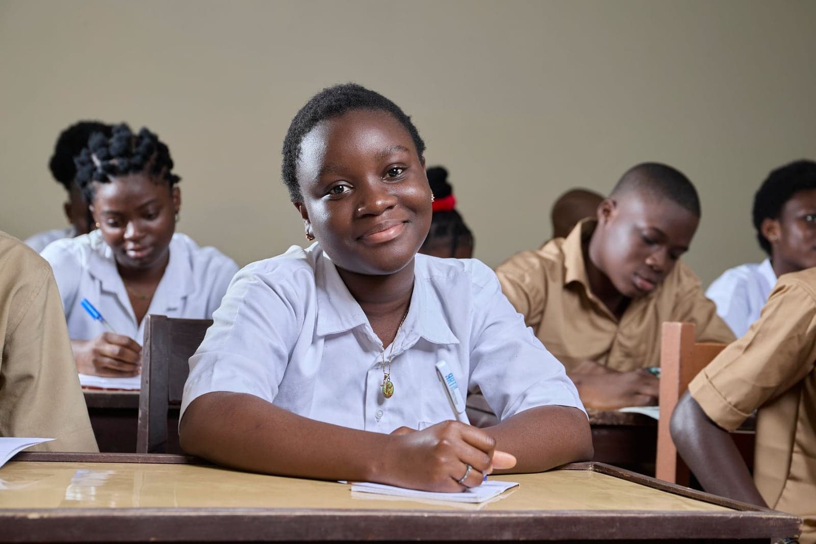 A secondary school student smiling at her desk, pen in hand, classmates working behind her.
