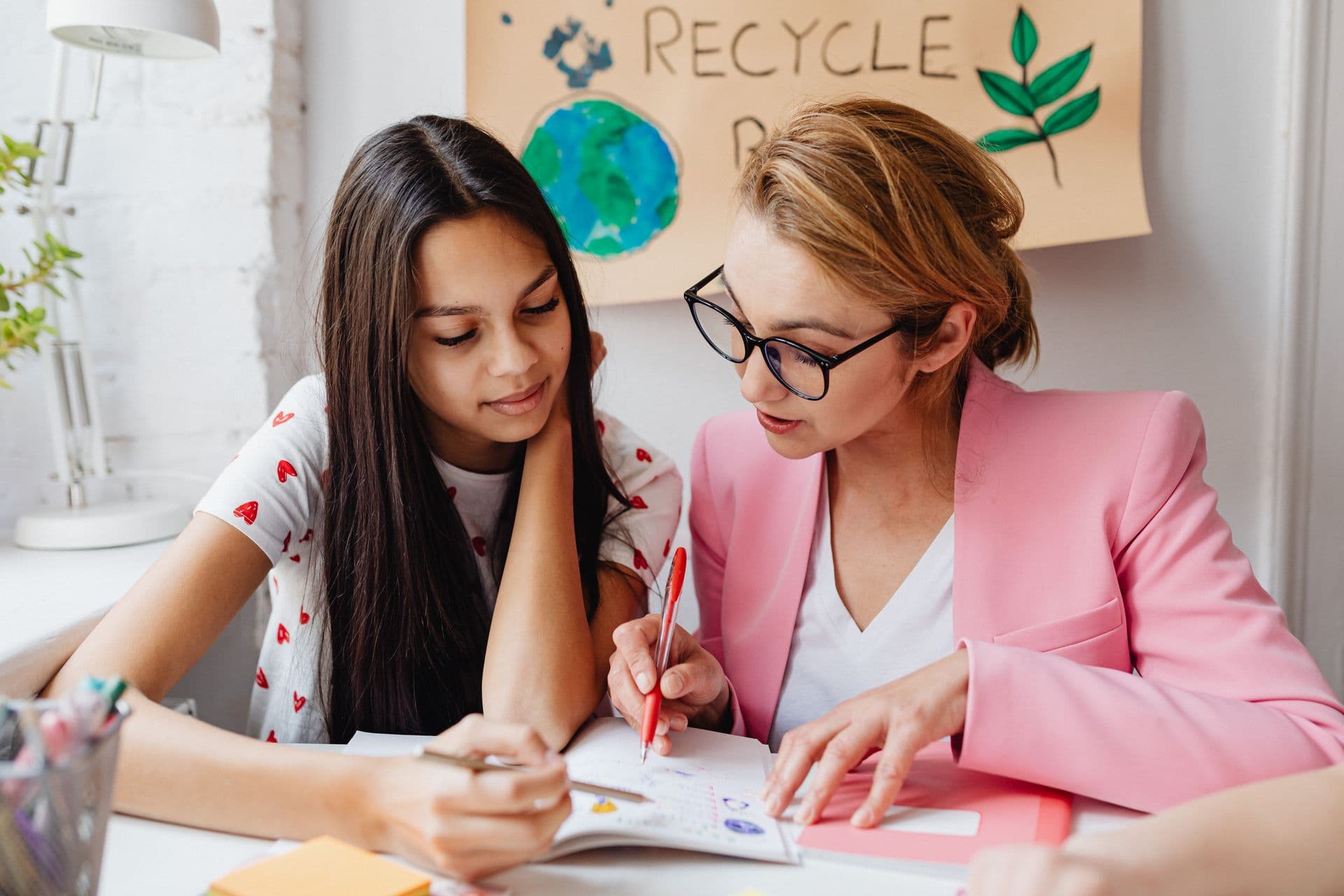 A teacher helping a student with her work beside a recycling poster.